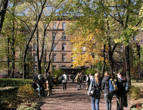 Zdjęcie budynku uczelnie za zielonymi drzewami i ścieżką, po której spacerują studenci.